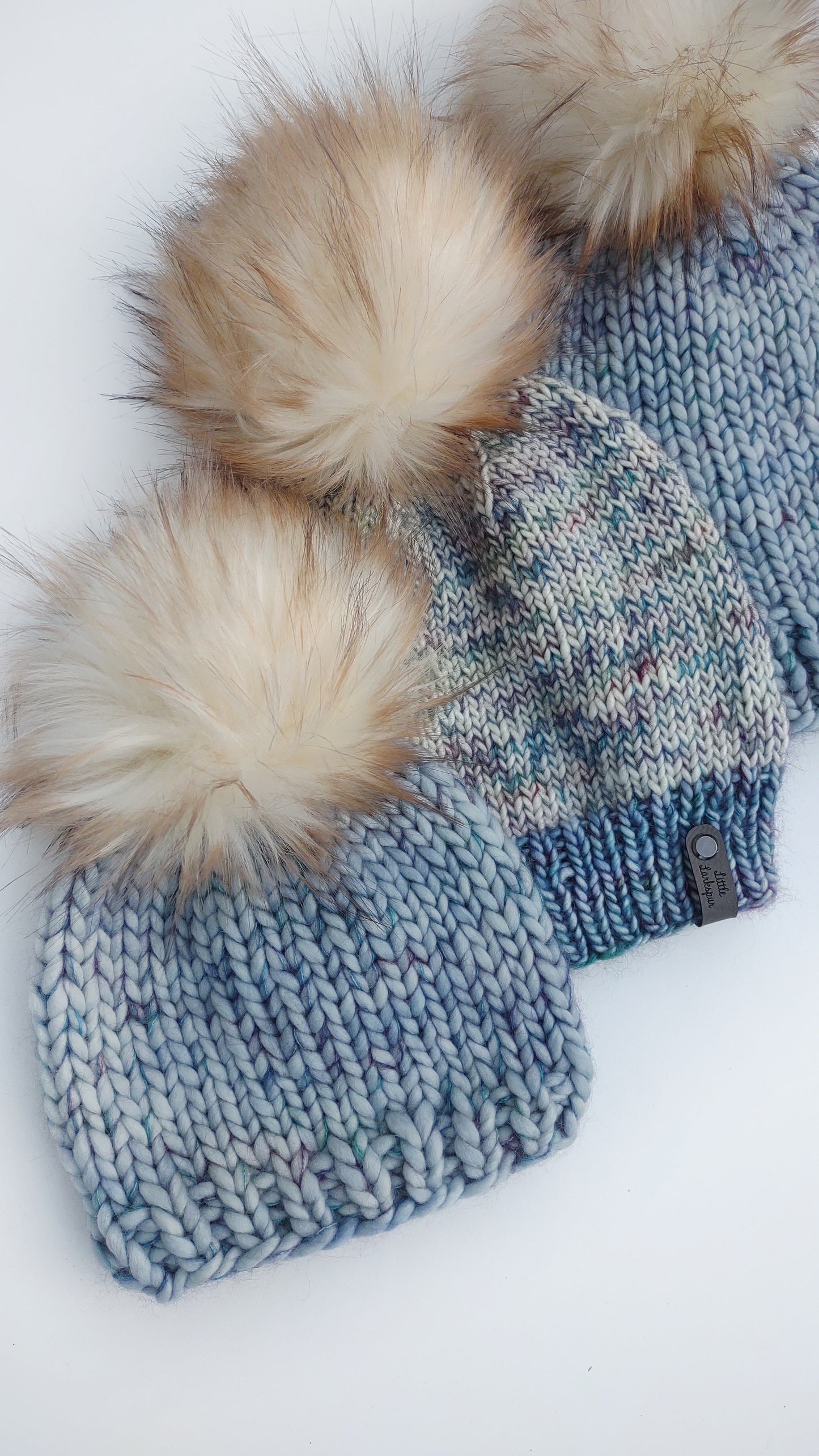 Flatlay of three blue hats with big fluffy white faux fur pom poms